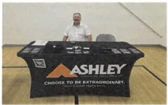 A man sitting at a table with promotional items and the text Ashley. 