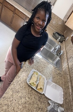 A young girl smiling for a picture with two tacos in a take out container.