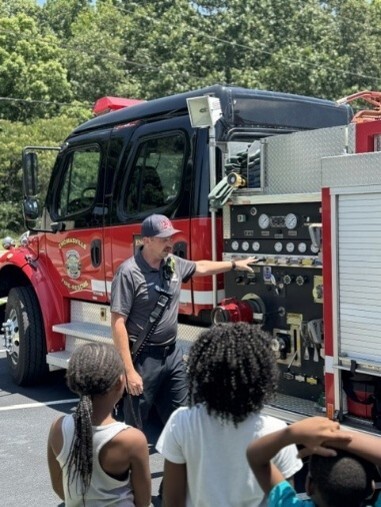 A man pointing to a part on a fire engine while kids watch.