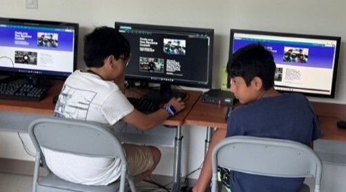 two boys sitting at a desk with computers.