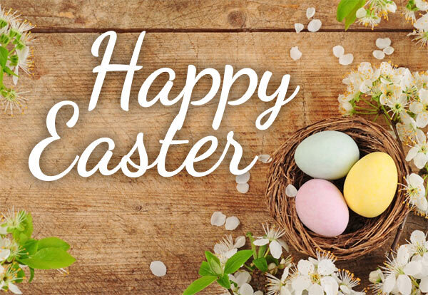 Happy Easter. Colorfully dyed eggs sit in a woven basket surrounded by flowers on a wooden table.